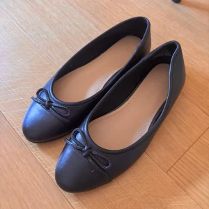 Svarta ballerinaskor med rosett från H&M premium - Säljer ett par klassiska svarta ballerinaskor från H&M Premium. Skorna har en rund tå och en söt liten rosett framtill. Perfekta för dig som vill ha en enkel och stilren sko till vardags. Tillverkade i syntetmaterial.