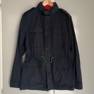 Mörkblå parkajacka med gröna och röda detaljer - Snygg mörkblå parkajacka med gröna och röd dragsko i midjan och röda detaljer vid knapparna. Jackan har fyra fickor framtill, axelklaffar och hög krage. Storlek Medium från Ganso. Blekare än ny. 
