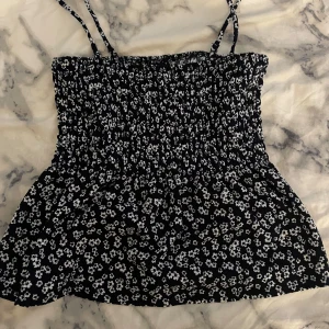 Svart blommig smockad linnetopp - Säljer en svart smockad linnetopp med vita små blommor och tunna avtagbara axelband. Toppen har en stretchig passform och är perfekt för varma dagar. Söt och enkel att matcha till olika outfits.