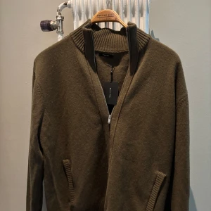 Olivgrön stickad tröja med dragkedja från Massimo Dutti - Säljer en olivgrön stickad tröja från Massimo Dutti med hög krage och dragkedja. Tröjan har långa ärmar och ribbade muddar vid ärmslut och nederkant. Perfekt för lager-på-lager och stilren look.