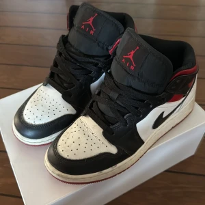 Nike Air Jordan 1 Mid svart/vit/röd - Säljer ett par Nike Air Jordan 1 Mid i klassisk svart, vit och röd färg. Skorna har svart swoosh, svarta snören och röd detalj vid ankeln. Ikonisk Jordan-logga på sidan och tungan. Perfekta för dig som gillar streetwear och sneakerskultur.