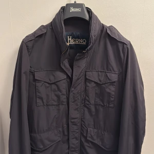 Herno jacka - Säljer en jacka från Herno med fyra fickor framtill och hög krage. Jackan har dragkedja och knappar, samt en stilren och enkel design. Perfekt för dig som vill ha en lätt och snygg jacka till vardags.