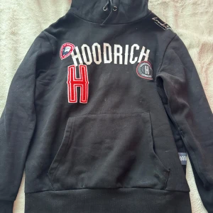 Svart hoodie från Hoodrich med broderade detaljer - Svart hoodie från Hoodrich med broderade märken och text på bröstet. Stor röd bokstav H och vita detaljer. Huva med dragsko och stor magficka framtill. Perfekt för en streetwear-look.