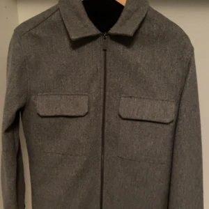 Grå overshirt med dragkedja - Säljer en stilren grå overshirt med dragkedja framtill och två bröstfickor med lock. Jackan har klassisk krage och lång ärm, perfekt för lager-på-lager under höst och vår. Priset kan diskuteras 