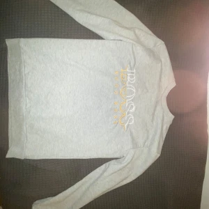 Grå långärmad tröja från Hugo Boss - Säljer en stilren grå långärmad tröja från Hugo Boss med vit och gul logotyp på bröstet. Perfekt för en avslappnad och snygg look. Klassisk passform och rund halsringning.