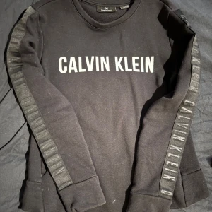 Svart långärmad tröja från Calvin Klein - Svart långärmad tröja från Calvin Klein Performance med stor vit logga framtill och logotyp längs ärmarna. Tröjan har en klassisk rund halsringning och är tillverkad i mjukt material, samt har två ”dolda” öppna fickor på sidorna. Aldrig använd och är kolsvart i färgen, bilden är lite missvisande i färgen. 
