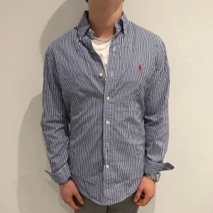 Blåvit randig skjorta från Polo Ralph Lauren - Säljer en klassisk blåvit randig skjorta från Polo Ralph Lauren i modellen Custom Fit. Skjortan har långa ärmar, knappar framtill och en röd broderad logga på bröstet. Perfekt för en stilren och tidlös look. Modellen är ca 172cm.