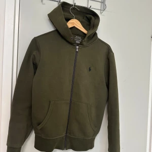 Olivgrön hoodie från Polo Ralph Lauren - Säljer en olivgrön hoodie från Polo Ralph Lauren med dragkedja framtill och två fickor. Tröjan har huva och en liten broderad logga på bröstet. Perfekt för en avslappnad stil. Köpte för 1700 säljer för 900 lite använt kan ner i pris passar både xs och s