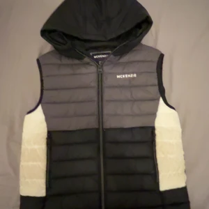Svart och grå dunväst med huva från McKenzie - Snygg dunväst från McKenzie i svart och grått med vita paneler på sidorna. Västen har en huva och dragkedja framtill samt diskret logga på bröstet. Perfekt för lager-på-lager och streetwear-stil.