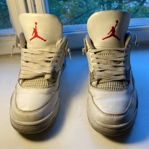 Vita Air Jordan 4 Oreo’s  - Säljer ett par klassiska vita Air Jordan 4 Retro med röda Jumpman-loggor på plös och sula. Skorna har snörning, meshpaneler på sidorna och en platt sula. Perfekta för dig som gillar streetwear och sneakers med ikonisk design. Ingen sula