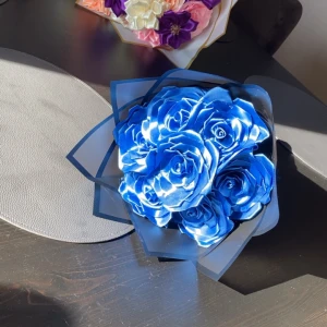 Bukett med blå konstgjorda rosor - Säljer en snygg bukett med blå konstgjorda rosor i satinliknande material. Blommorna är arrangerade i ett svart pappersomslag med en matchande blå rosett. Perfekt som present eller dekoration.