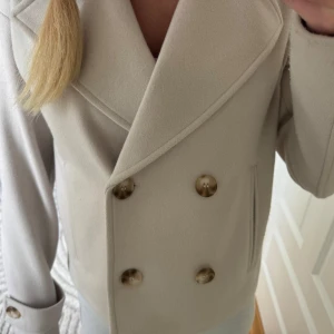 Beige kappa från Kappahl - Säljer en stilren beige kappa från Kappahl med dubbelknäppning och bred krage. Jackan har långa ärmar och dekorativa knappar vid ärmslut. Perfekt för dig som gillar klassisk och clean stil. Den är använd 1 gång.