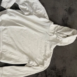 Vit hoodie - Säljer en klassisk vit hoodie med huva och dragsko. Perfekt för en avslappnad stil och enkel att matcha med det mesta. Mjuk och skön med långa ärmar och bred mudd vid ärmslut och nederkant.