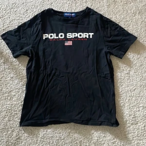 Svart t-shirt från Polo Sport Ralph Lauren - Säljer en svart t-shirt från Polo Sport Ralph Lauren med tryck i vitt och rött framtill samt en liten amerikansk flagga. Klassisk rund halsringning och normal passform. Perfekt för en sportig och stilren look.