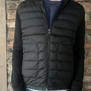 Svart pufferjacka från Moncler - Snygg svart pufferjacka från Moncler med stickade ärmar och quiltad front. Jackan har dragkedja framtill och Moncler-logga på ärmen. Perfekt för dig som vill ha en stilren och sportig look. Den är i storlek M och är köpt på nk i Göteborg. Den är i riktigt bra skick. Har bara används fåtal gånger. Säljer den för att den har blivit för liten oxh jag är 180 passar bäst för nån som e 170-176. Kvitto kommer priv i dm skriv om du har några frågor!!