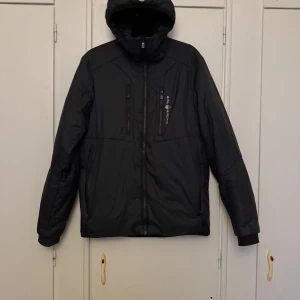 Svart jacka med huva från Sail Racing - Säljer en svart jacka från Sail Racing med huva och dragkedja framtill. Jackan har två bröstfickor med dragkedja och en sportig look.
