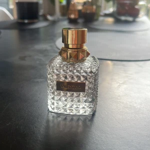 Valentino parfymflaska Donna 50 ml - Säljer en lyxig parfym från Valentino i en transparent, mönstrad glasflaska med guldfärgat lock och detaljer. Flaskan har en fyrkantig form och en elegant etikett framtill. Perfekt för dig som gillar exklusiva dofter och snygg design. Nypris 1300kr. Endast använd väldigt lite. 