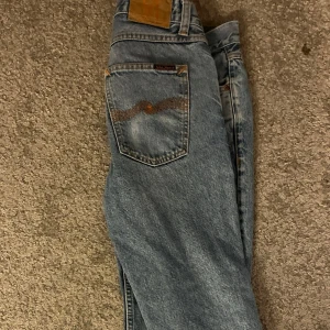 Ljusblå jeans från Nudie Jeans - Säljer ett par klassiska ljusblå jeans från Nudie Jeans med raka ben och femficksdesign. Jeansen har Nudie’s signaturbroderi på bakfickorna och är tillverkade i slitstarkt denim. Perfekta för en avslappnad stil. Storlek:30/30