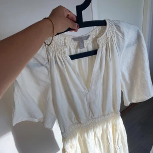 Vit omlottklänning med v-ringning - Säljer en luftig vit klänning med v-ringning och korta vida ärmar. Klänningen har resår i midjan och ett lätt glansigt material som ger en elegant känsla. Perfekt för varma dagar och somriga tillfällen.