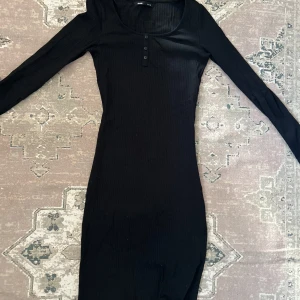 Svart långärmad klänning från Sinsay - Svart, figurnära klänning från Sinsay med ribbad struktur och långa ärmar. Klänningen har rund halsringning och tre knappar framtill för en enkel men stilren look. Perfekt för dig som gillar minimalistisk stil.