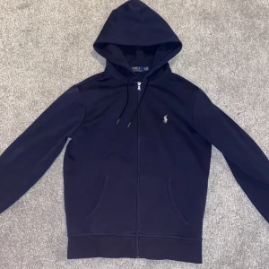 Marinblå hoodie från Polo Ralph Lauren - Säljer en stilren marinblå hoodie från Polo Ralph Lauren med dragkedja och det klassiska broderade logotypen på bröstet. Perfekt för en avslappnad look. Den har långa ärmar och en praktisk huva med dragsko. Har knappast använt den så den är i nästan nytt skick. Nypris 2000kr