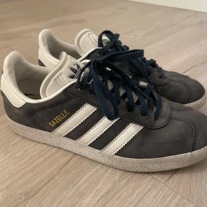 Adidas Gazelle sneakers i blått/grått och vitt - Säljer ett par klassiska Adidas Gazelle sneakers i grå/blå mocka med vita detaljer och mörkblå snörning. Skorna har de ikoniska tre ränderna på sidan och guldfärgad Gazelle-text. Perfekta för en sportig och stilren look.