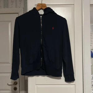Mörkblå hoodie från Polo Ralph Lauren - Mörkblå hoodie från Polo Ralph Lauren med dragkedja och huva. Tröjan har två fickor framtill och en röd broderad logga på bröstet. Klassisk och stilren modell som passar perfekt till vardags.