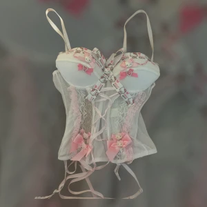 Vit korsett med Hello Kitty-detaljer och rosetter - Supersöt vit korsett med spets, rosa rosetter och Hello Kitty-band. Dekorativa knytband framtill och spetsdetaljer längs sidorna. Perfekt för dig som gillar gulliga och unika plagg!