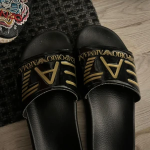 Svarta badsandaler från Emporio Armani EA7 - Svarta badsandaler från Emporio Armani EA7 med guldiga detaljer och logga över foten. Sköna att ha på sig och perfekta för sommaren eller till stranden.