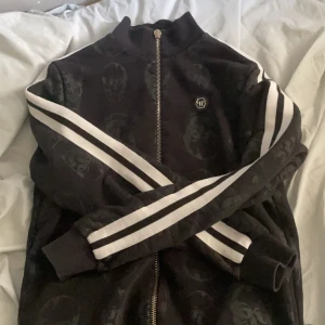 Svart hoodie med dödskallemönster från Philipp Plein - Svart hoodie med subtilt dödskallemönster och vita ränder längs ärmarna. Jackan har dragkedja framtill och en liten logotyp på bröstet. Dragkedjan saknar delen man använder för att dra upp den, men annars är de inga fel :). Storlek S.
