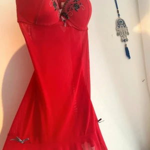 Vintage negligé topp mesh broderi 00s 2000s y2k - En vintage neglige från 00-talet i genomskinligt rött meshtyg med detaljer i svart spets och broderade rosor på kuporna. Modellen har en bygel med vadderade C-kupor och justerbara axelband, samt dekorativa rosetter i svart och silver. Nedtill är plagget utsmyckat med en liten volangkant. Tyget är stretchigt och lätt, vilket gör plagget följsamt mot kroppen. Märkt “Indulgence by Biatta”. Ingen storleksangivelse, men den passar ungefär som en M och C-kupa. Väldigt fint skick. 