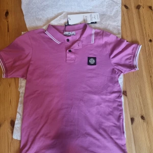 Rosa piké t-shirt från Stone Island - Säljer en rosa piké t-shirt från Stone Island med vit kant vid krage och ärmslut. Klassisk polokrage med två knappar och broderad logotyp på bröstet. Perfekt för dig som gillar stilrena märkeskläder. Helt ny aldrig använd. 