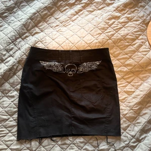 Svart kortkjol med döskalle och vingar - Säljer en svart kortkjol med coolt döskallemotiv och vingar i strass på baksidan. Kjolen har klassisk femficksdesign, knapp framtill och råa kanter nertill. Perfekt för dig som gillar edgy stil och detaljer som sticker ut.
