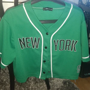 Grön baseballinspirerad tröja  - Säljer en grön baseballtröja från FB STER med texten 'NEW YORK' framtill och siffran 92 på ärmen. Tröjan har korta ärmar, knappar framtill och vita detaljer längs kanterna. Perfekt för en sportig look!