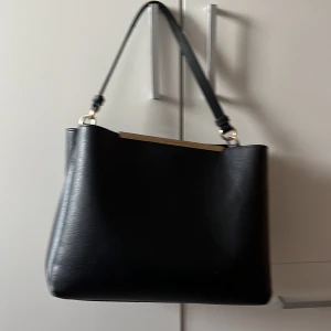 Svart axelväska i skinn - Snygg svart axelväska i skinn med stilren design och guldfärgade metalldetaljer. Väskan har en enkel, rymlig form och ett avtagbart axelband. Perfekt för dig som vill ha en klassisk accessoar med plats för allt du behöver.