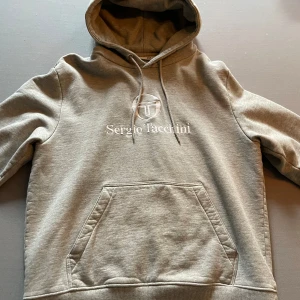 Grå hoodie från Sergio Tacchini - Säljer en stilren grå hoodie från Sergio Tacchini med broderad logga och känguruficka framtill. Mjuk insida och justerbar huva med dragsko. Perfekt för en avslappnad look.