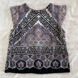 Mönstrad blus från Oasis - Säljer en snygg blus från Oasis med orientaliskt inspirerat mönster i svart, rosa, blått och vitt. Blusen har korta ärmar och en liten knappdetalj i nacken. Perfekt för dig som gillar färg och mönster! Prislappen sitter löst men annars är den i mycket bra skick! 