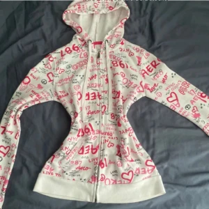  Aero kofta - Säljer en vit kofta från Aeromed färgglatt tryck i rött och svart. Hoodien har dragkedja framtill och en mysig huva. Perfekt för en avslappnad stil!