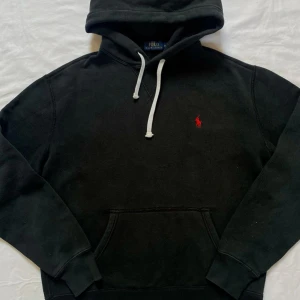 Svart hoodie från Ralph Lauren - Säljer en klassisk svart hoodie från Ralph Lauren med det ikoniska röda logotypbroderiet på bröstet. Tröjan har en känguruficka och justerbar huva med vita snören. Perfekt för en avslappnad stil.