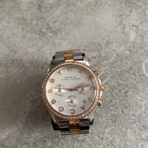 Tvåfärgad klocka från Marc Jacobs - Snygg klocka från Marc Jacobs med en tvåfärgad länk i silver och guld. Urtavlan är vit med guldiga detaljer och bokstäver runt kanten. ORD pris 2800kr