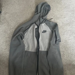 Grå hoodie från Nike - Säljer en grå hoodie från Nike med dragkedja och huva. Den har en klassisk design med Nike-logga på bröstet. Perfekt för en avslappnad stil.