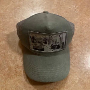 Grå keps  - Snygg grön keps med nätpaneler på sidorna och ett tryck framtill som föreställer en stadsmiljö. Perfekt för en avslappnad stil.