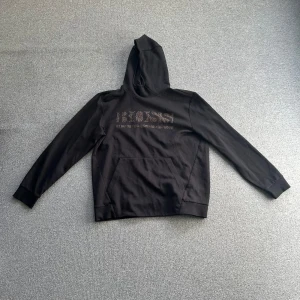 Svart hoodie från Hugo Boss - Snygg svart hoodie från Hugo Boss med logga på bröstet. Tröjan har en klassisk design med huva och en stor ficka framtill. Perfekt för en stilren och bekväm look. Den är i XL fast den är liten i storleken och passar bäst på folk som har L.