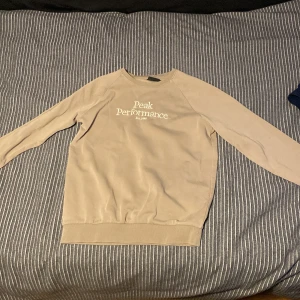 Beige tröja från Peak Performance - Säljer en beige tröja från Peak Performance med rund hals och långa ärmar. Tröjan har en enkel design med logotypen tryckt på framsidan. Perfekt för en avslappnad stil.