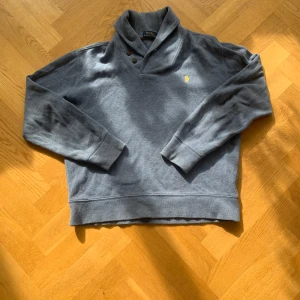 Grå tröja från Ralph Lauren - Säljer en stilren grå tröja från Ralph Lauren med en guldig broderad logga på bröstet. Tröjan har en unik krage med två knappar och långa ärmar. Perfekt för en avslappnad look.