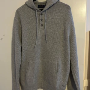Grå stickad cashmere hoodie från Polo Ralph Lauren - (Aldrig använd) Säljer en stilren grå stickad hoodie från Polo Ralph Lauren. Tröjan har en knappslå med tre knappar och en praktisk magficka. Perfekt för kyliga dagar med sin mjuka och bekväma passform.