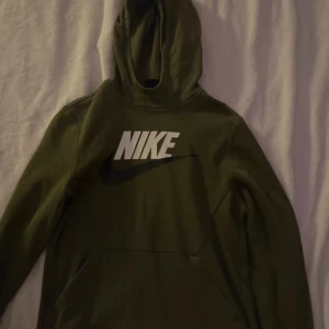 Olivgrön hoodie från Nike - Säljer en olivgrön hoodie från Nike med stor vit logga på bröstet. Tröjan har en klassisk design med huva och en stor magficka framtill. Perfekt för en avslappnad stil.