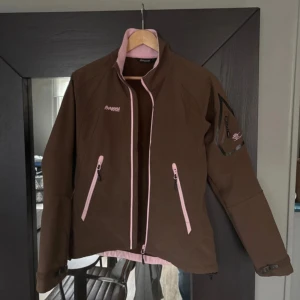 Brun funktions / skal jacka från Bergans Stl S - Säljer en brun jacka från Bergans med rosa detaljer och dragkedjor. Jackan har en praktisk ficka på ärmen och är perfekt för utomhusaktiviteter. Den har en normal passform och är långärmad. Perfekt för höst och vår! Modell: 1553 Stamsund 2 Lady   Stl S  Obs spännena vid händerna har släppt men annars är jackan jätte fin!