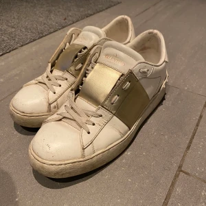 Valentino Garavani sneakers i vitt och guld - Valentino Garavani sneakers i vitt skinn med guldiga detaljer. Skorna har en unik design med breda guldiga band över snörningen och nitar på hälen. Box, extra skosnören och äkthetsintyg medföljer