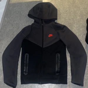Svart hoodie från Nike - Säljer en svart hoodie från Nike med röd logga på bröstet. Den har en dragkedja framtill och två fickor med dragkedjor. Perfekt för en sportig look eller avslappnad stil. Huvan ger extra skydd och komfort. Den är i perfekt skick. Det ända problemet är att dragthedian är lite trasig.